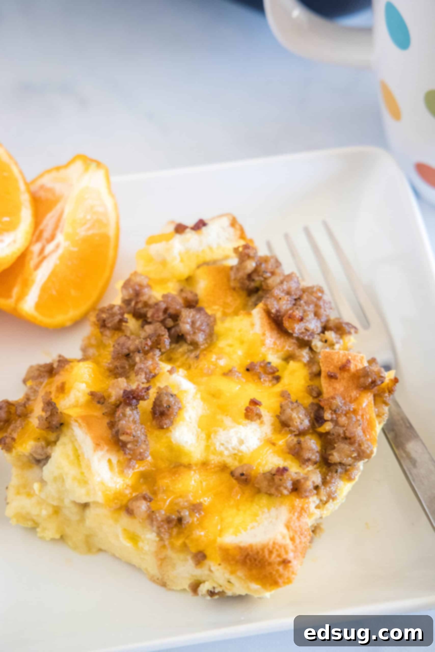A serving of sourdough breakfast casserole next to orange slices and a fork on a white square plate.