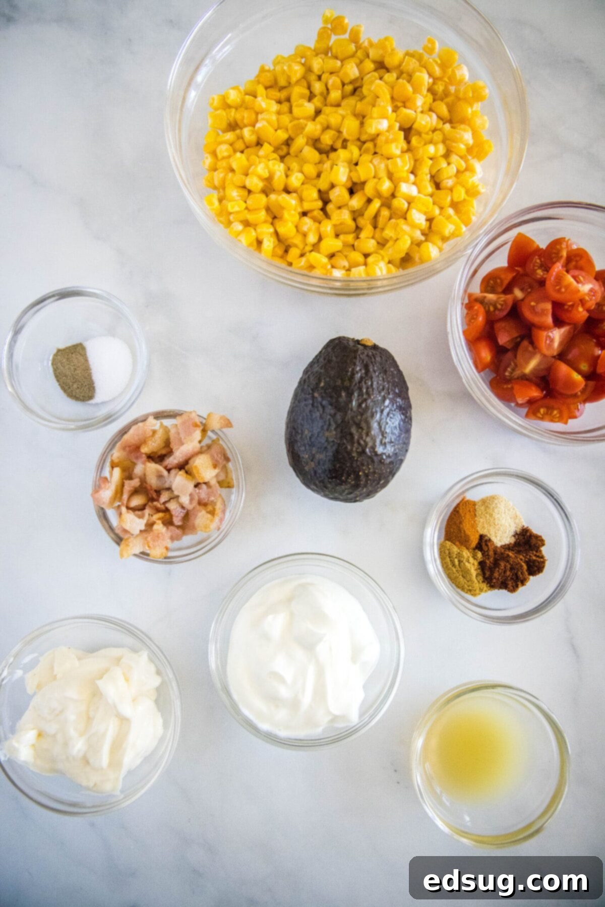 Overhead view of the ingredients needed for southwestern corn salad: a bowl of corn, a bowl of quartered cherry tomatoes, a bowl of cooked bacon crumbles, a bowl of salt and pepper, a bowl of greek yogurt, a bowl of mayo, a bowl of lime juice, a bowl of chili powder, cumin, garlic powder, and black pepper, and an avocado