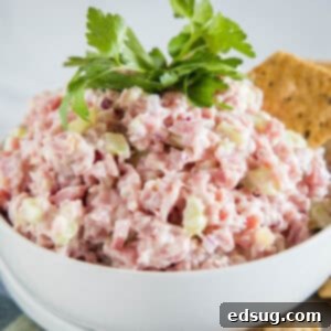Ham salad in a white bowl garnished with a cracker and fresh parsley leaves.