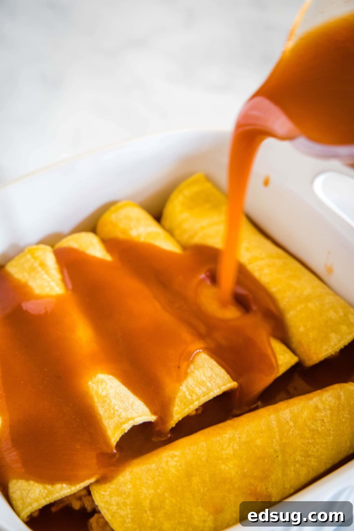 Enchilada sauce being generously poured over a tray of rolled chicken enchiladas in a baking dish, before baking.