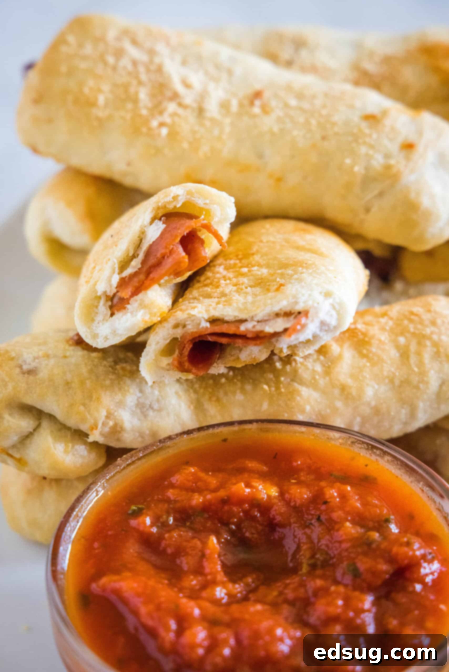 Pepperoni rolls stacked behind a bowl of marinara sauce, with one roll broken in half showing the melted cheese and pepperoni inside.