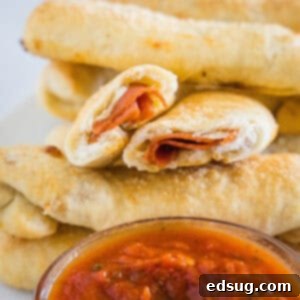 Homemade pepperoni rolls stacked behind a bowl of marinara sauce, with one roll broken in half revealing the melted cheese and pepperoni inside.