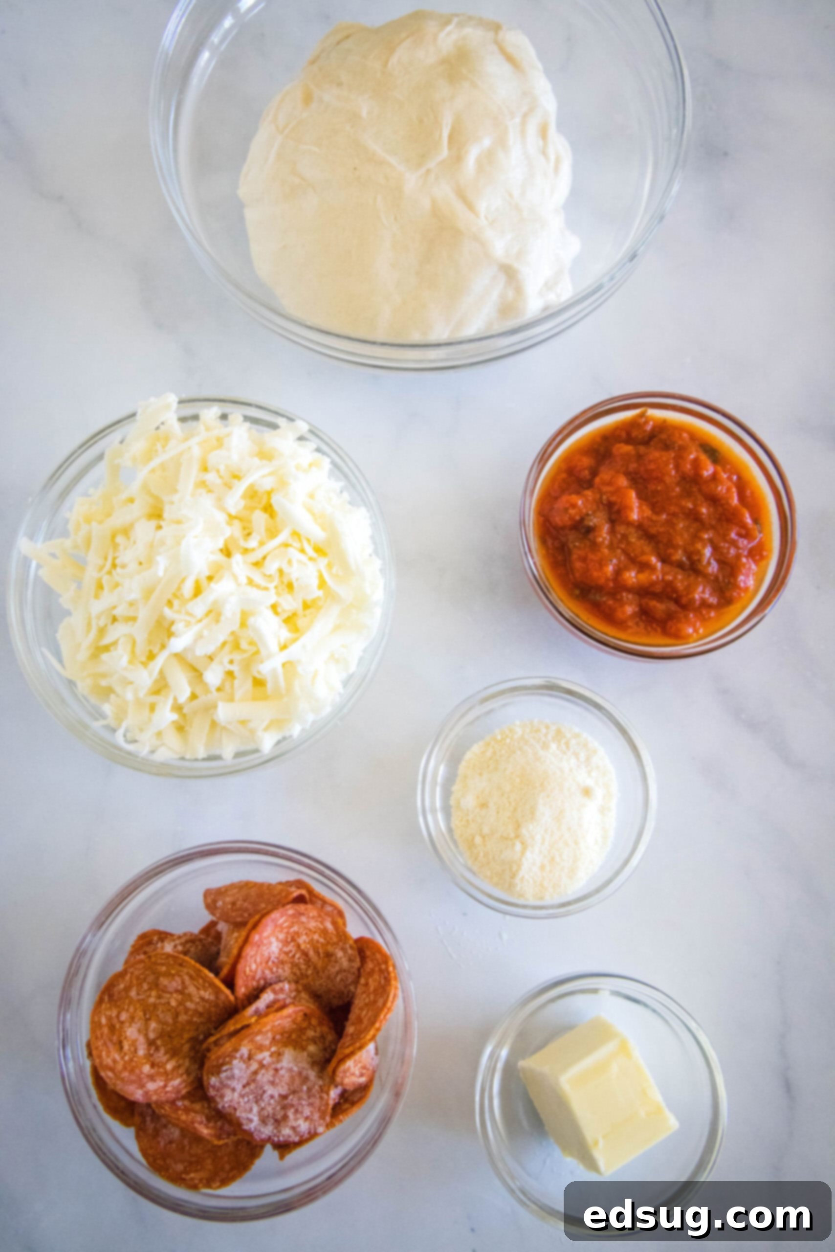 Various ingredients for pepperoni rolls laid out: pizza dough, shredded mozzarella, pepperoni slices, butter, and parmesan cheese.