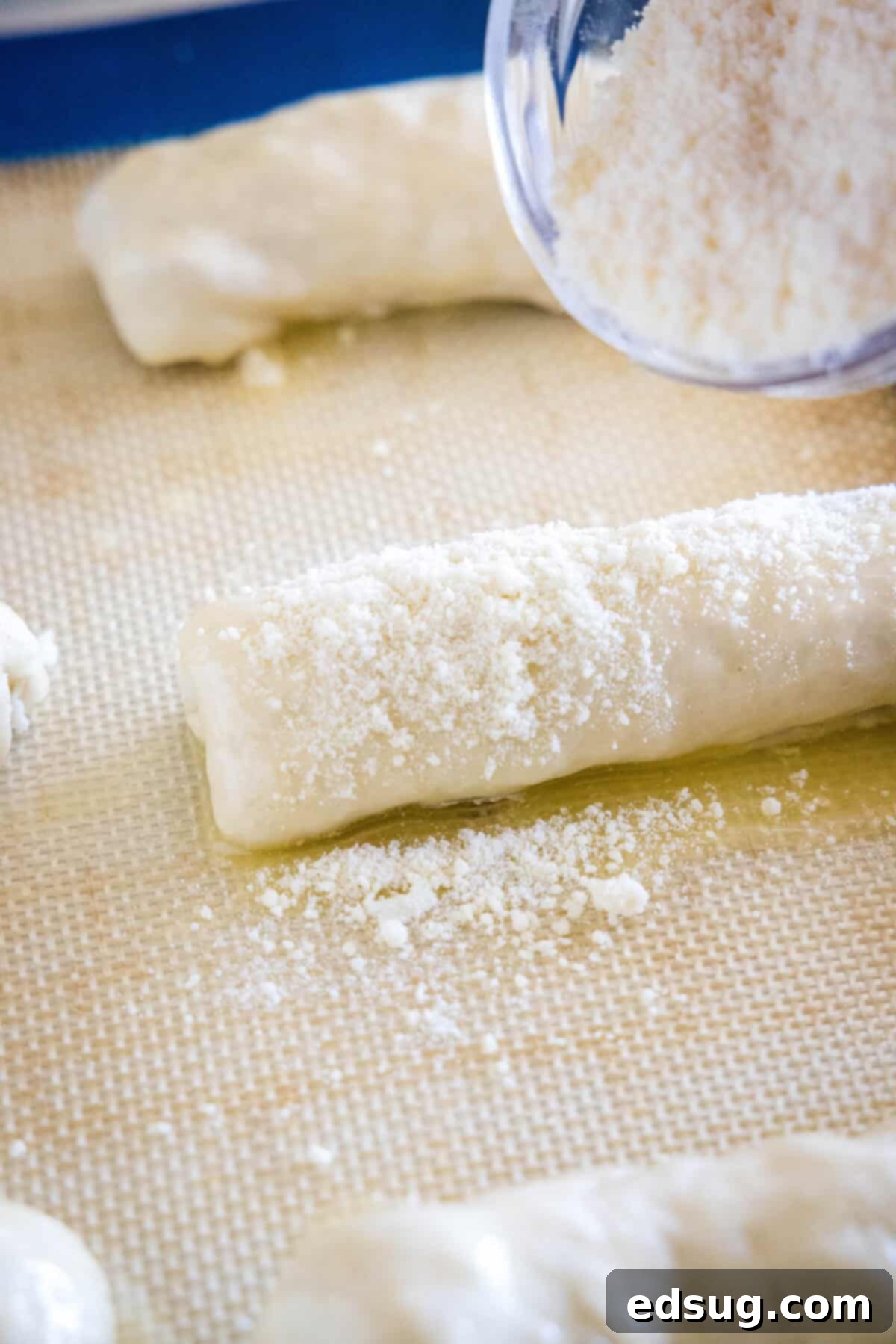 Grated parmesan being sprinkled from a small bowl over top prepared pepperoni rolls arranged on a lined baking sheet.