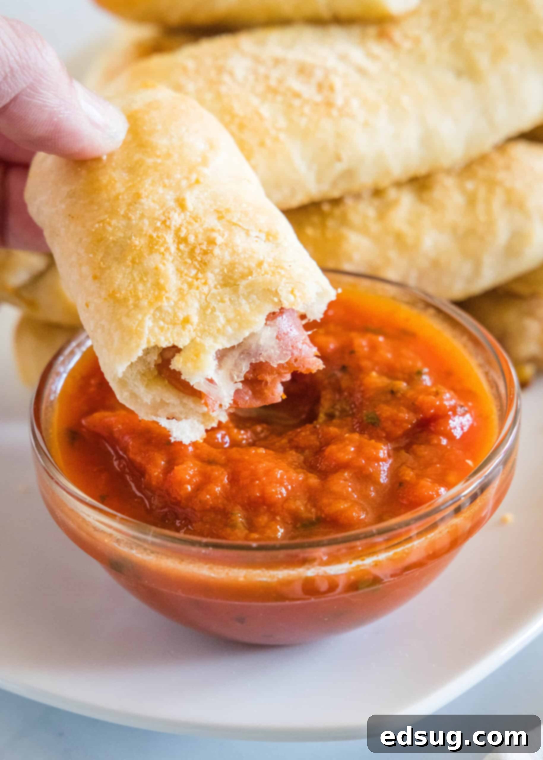 A hand holding one half of a pepperoni roll above a small bowl of marinara sauce, showing the melted cheese inside, with more rolls in the background.