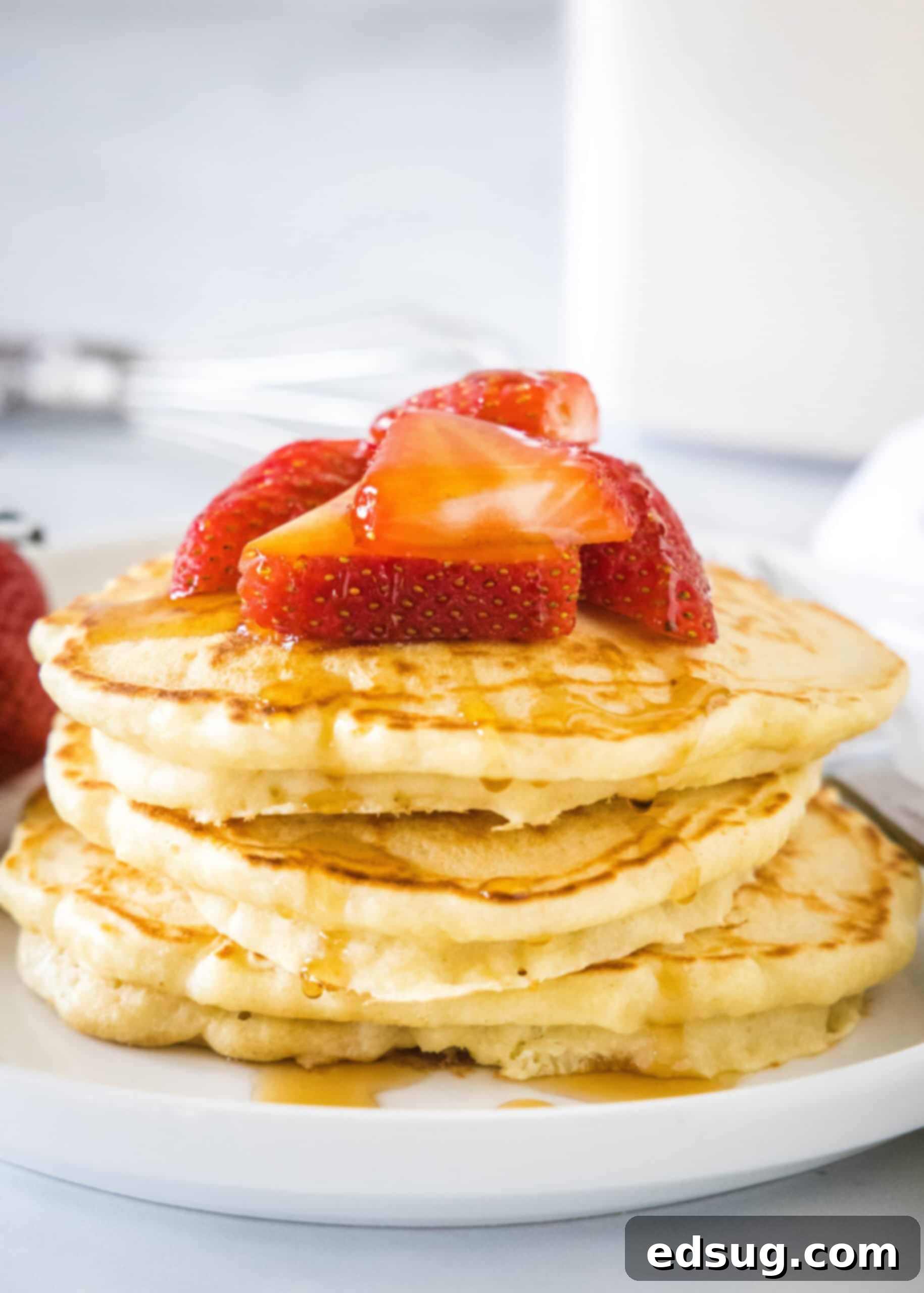 A stack of homemade pancakes topped with fresh strawberries and syrup on a white plate. They are golden brown and look incredibly fluffy.
