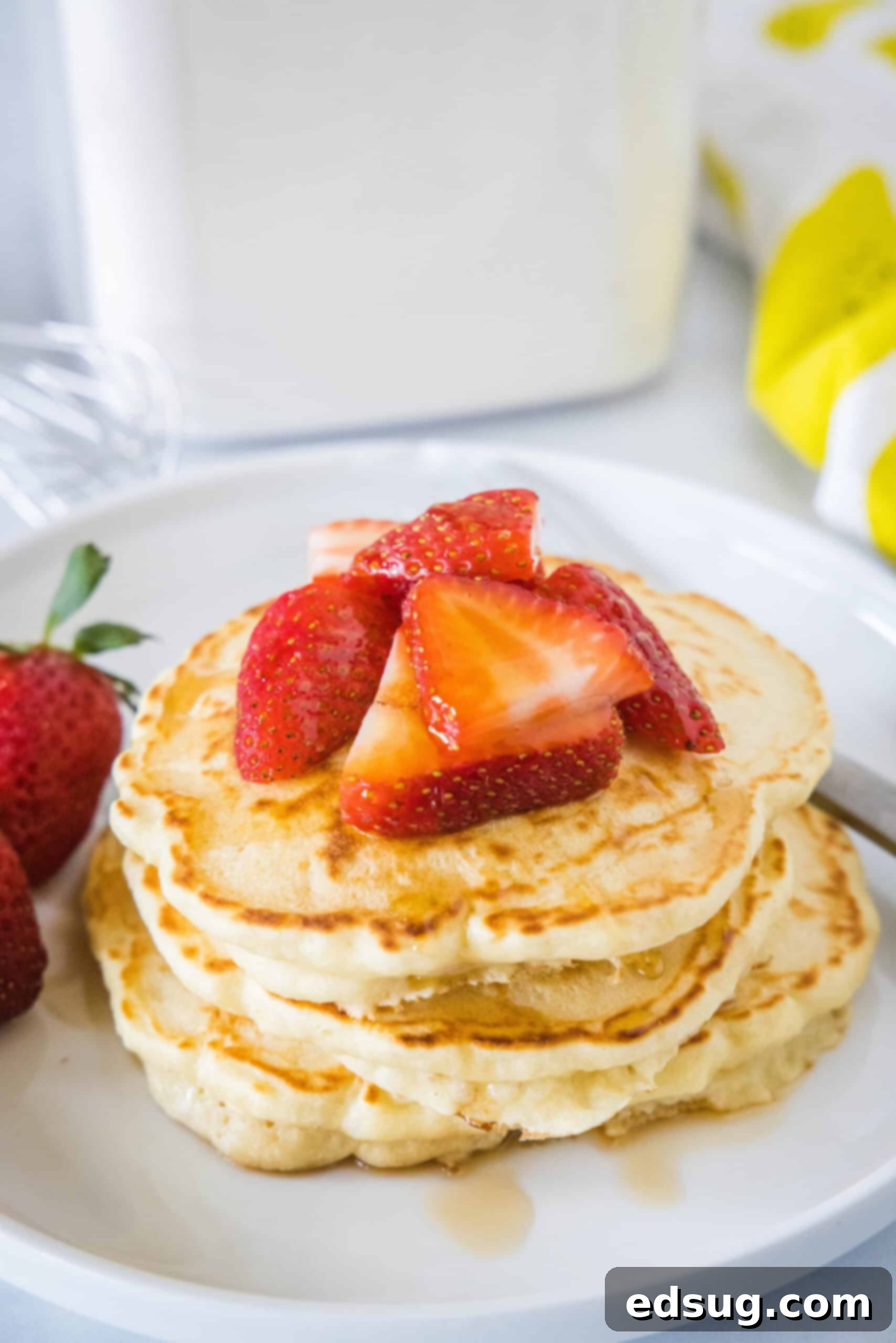 A beautifully presented stack of fluffy homemade pancakes, generously drizzled with maple syrup and adorned with fresh, vibrant strawberries on a white plate.