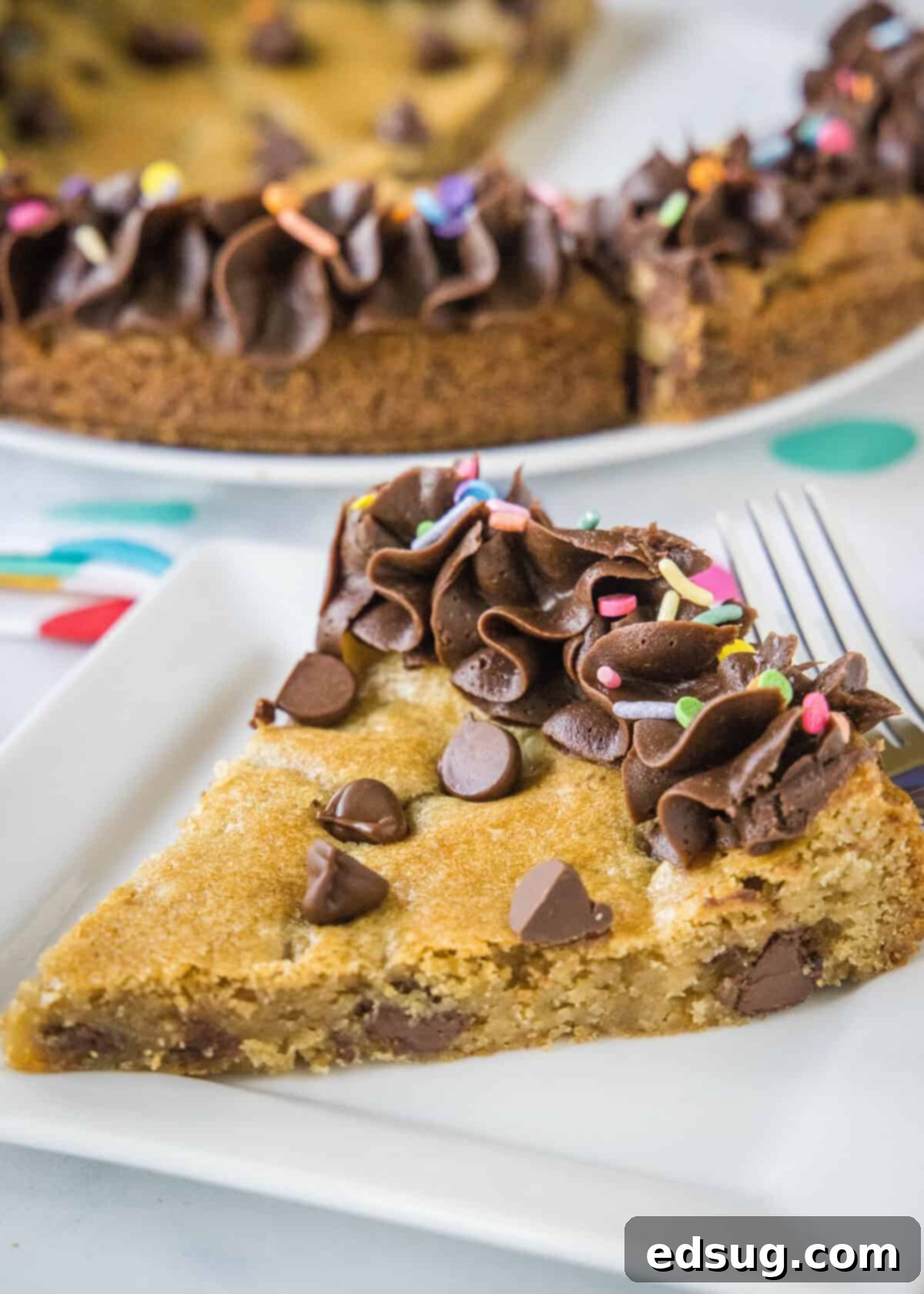 A generous slice of chewy chocolate chip cookie cake, topped with creamy frosting, served on a white plate.