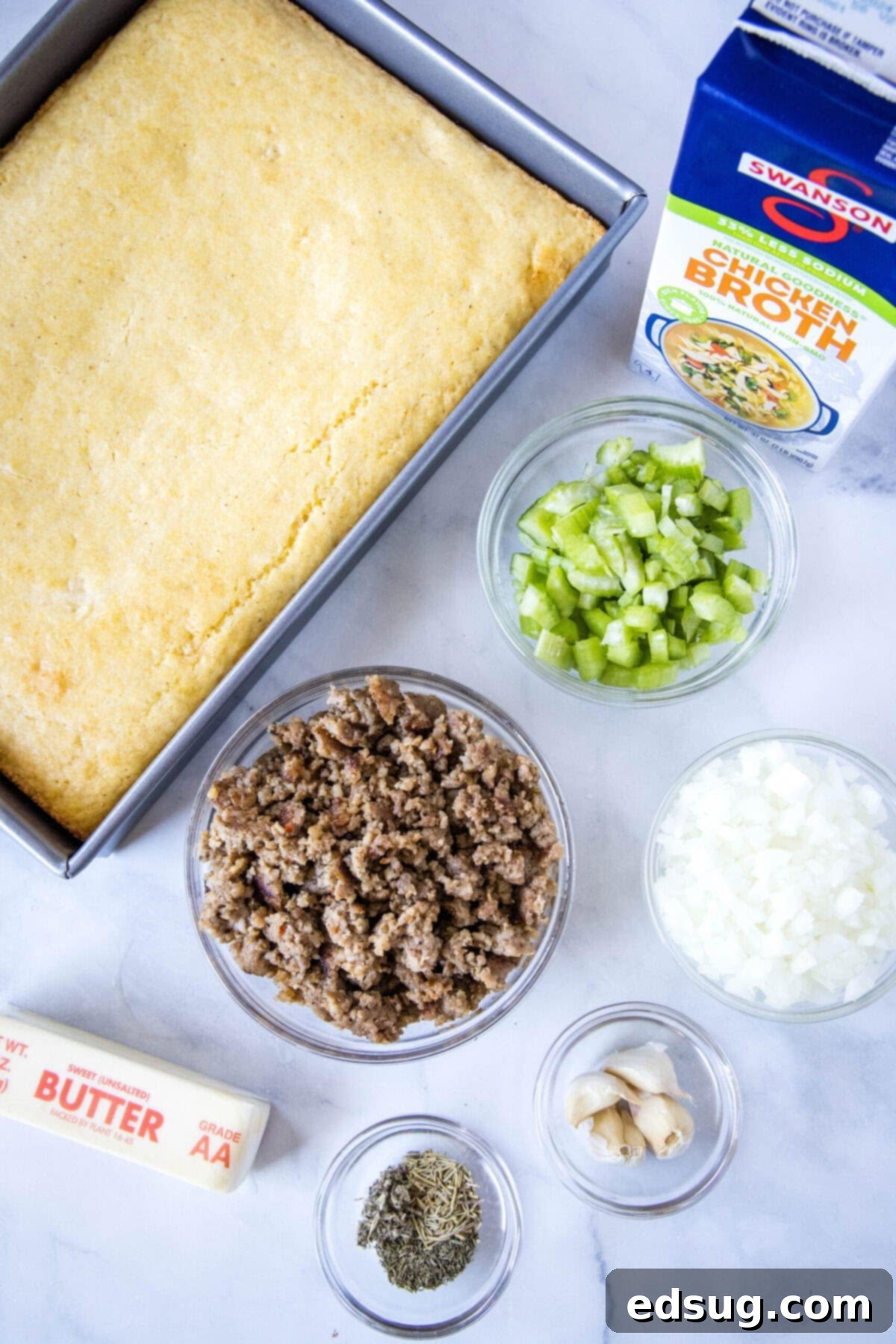 Overhead view of the ingredients needed for cornbread stuffing: a dish of cornbread, a bowl of sausage meat, a bowl of onions, a bowl of celery, a stick of butter, a bottle of chicken stock, a bowl of garlic, and a bowl of dried spices