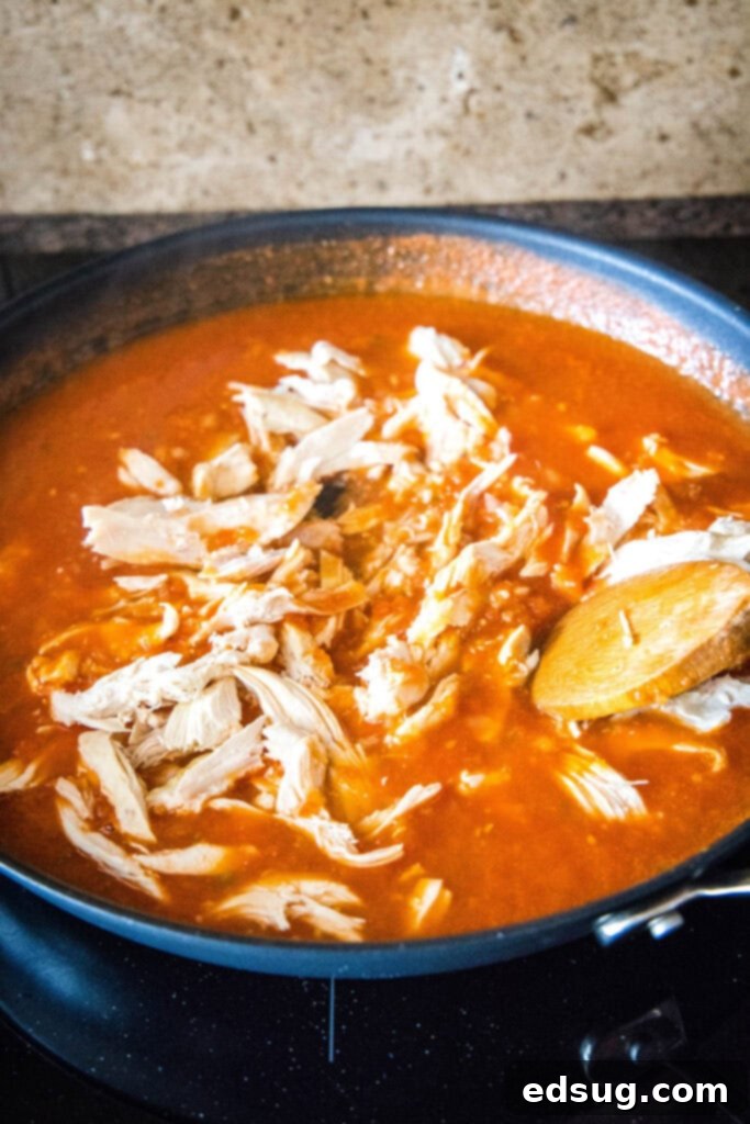 Shredded chicken being added to a simmering, rich red tomato sauce in a large skillet on the stovetop.