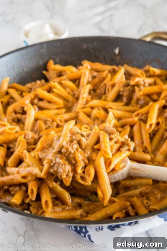 Hearty penne pasta with ground beef, vibrant spices, and gooey cheddar cheese bubbling in a cast iron skillet, garnished with fresh herbs and ready to serve.