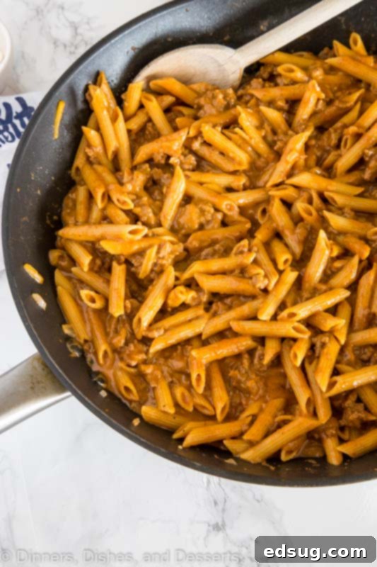 Uncooked short-cut pasta being added to a large pot with browned ground beef, onions, and spices, before liquid is added to begin simmering.