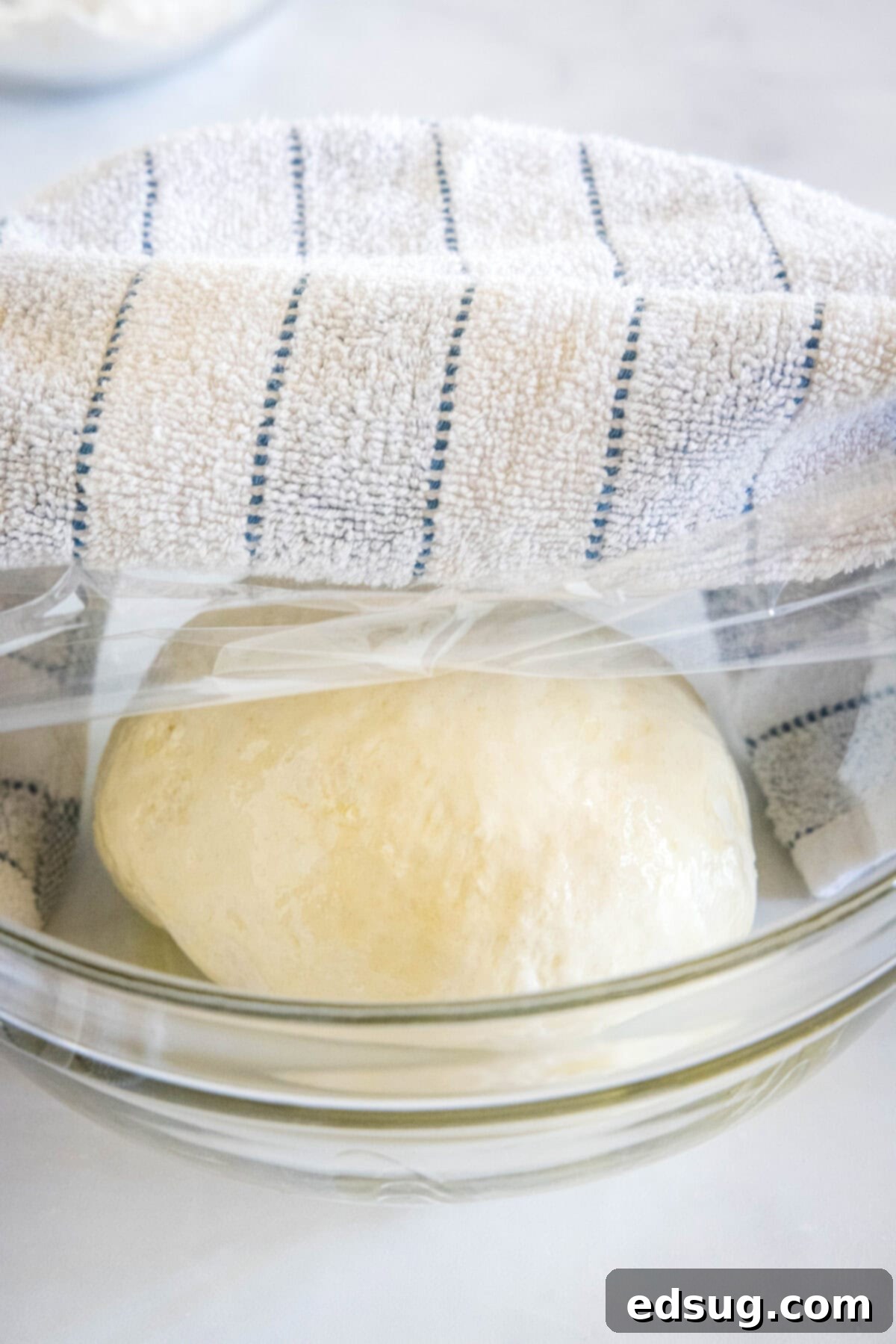 Crafting Perfect Pita 7 A perfectly smooth ball of pita bread dough rising in a glass bowl, covered with plastic wrap and a dishcloth.