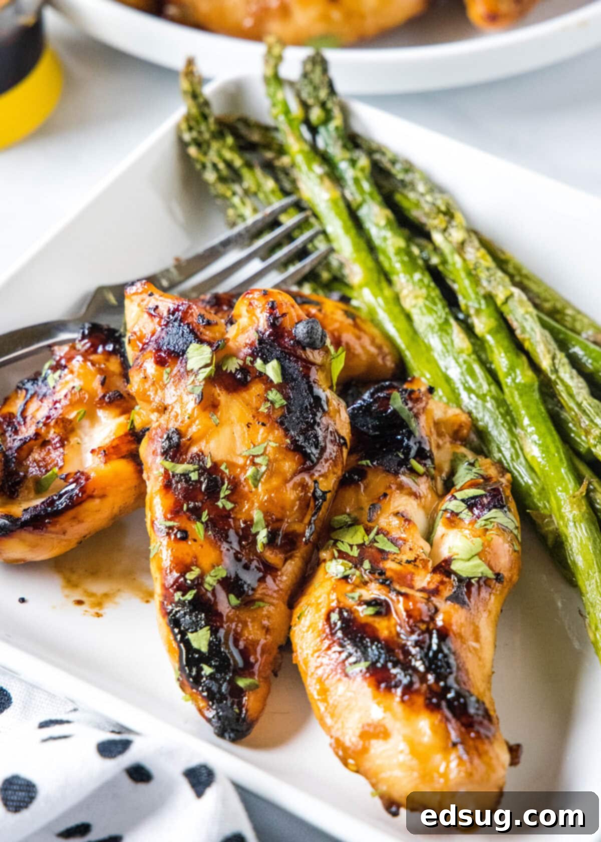 Sweet & Sticky Grilled Teriyaki Chicken 8 A serving of grilled teriyaki chicken on a square white plate, accompanied by a side of steamed asparagus and a fork, ready for a delicious meal.