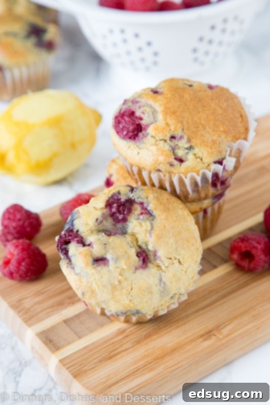 A close-up of a homemade lemon raspberry muffin with a golden crust and visible raspberries, surrounded by fresh lemons and berries on a wooden surface.