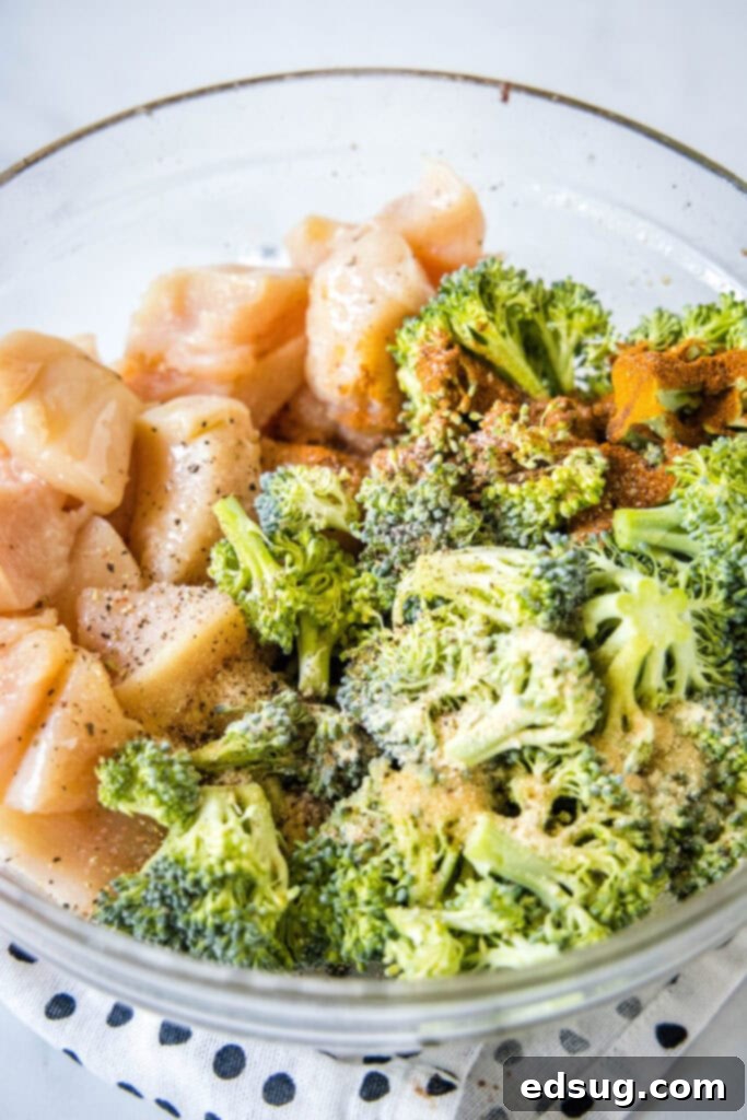A mixing bowl filled with raw broccoli florets and diced chicken, glistening with olive oil and coated in a vibrant blend of paprika, garlic powder, salt, and pepper.