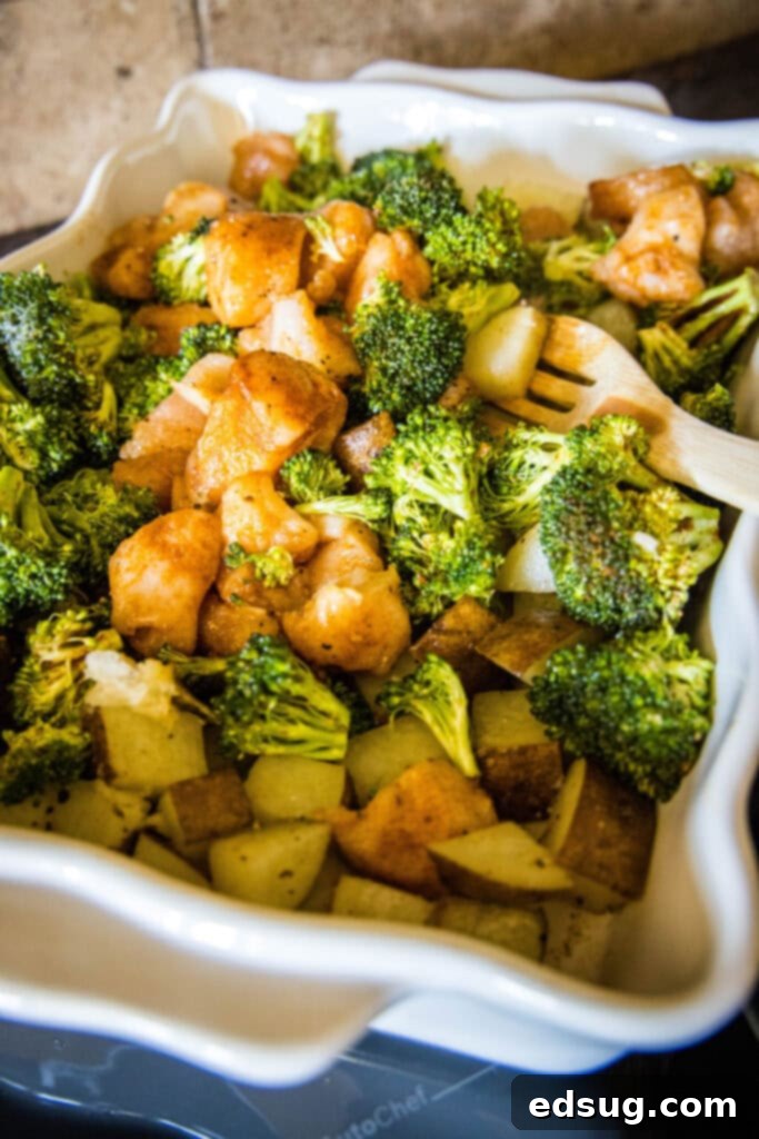 A 13x9-inch casserole dish containing a vibrant mixture of partially baked potatoes, tender broccoli, and succulent chicken pieces, with a wooden spoon stirring the ingredients to combine them before the final bake.