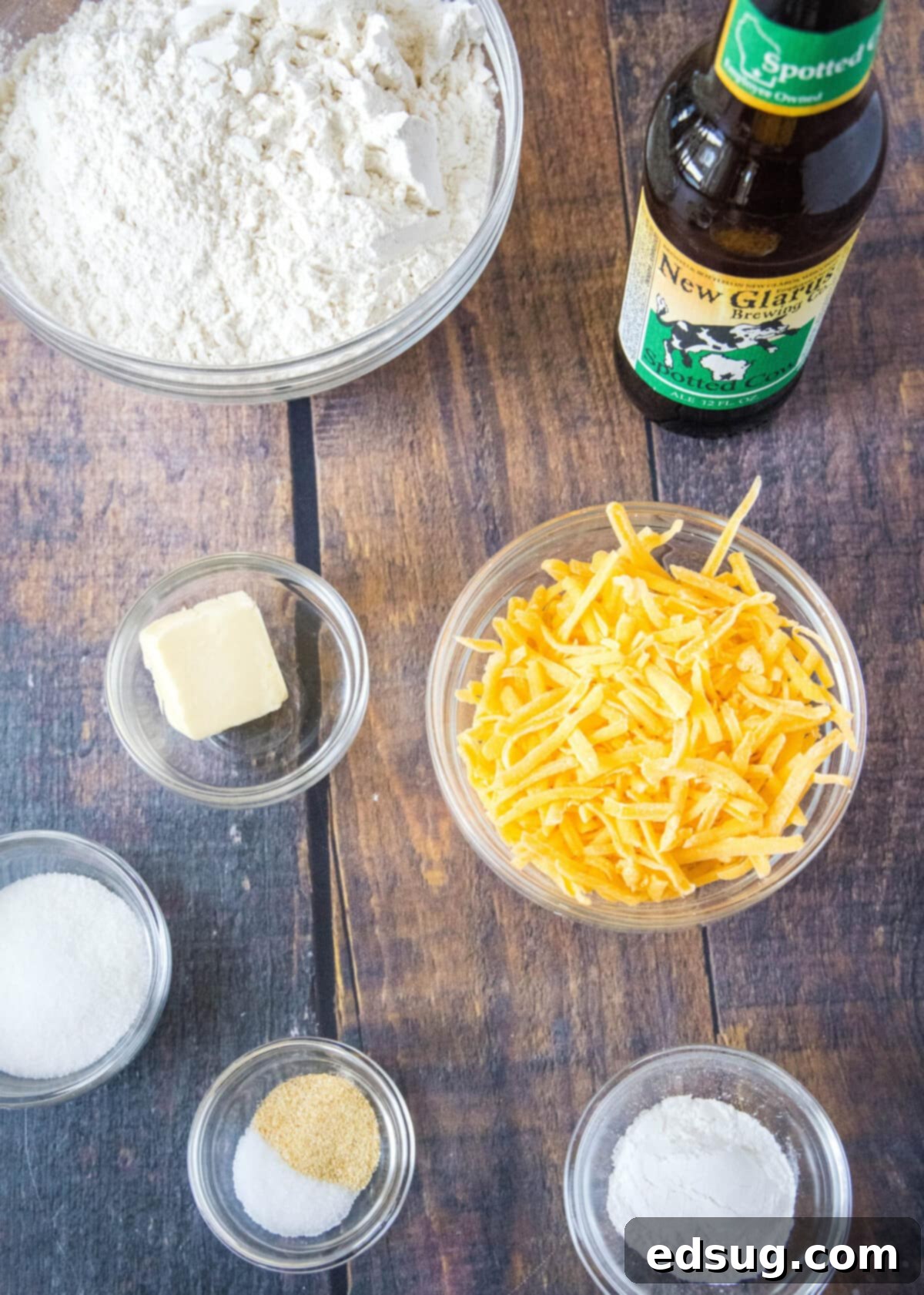 Overhead view of all the essential ingredients laid out meticulously for making homemade cheddar beer bread: a bowl of all-purpose flour, a bowl of shredded cheddar cheese, baking powder, granulated sugar, salt, garlic powder, a stick of butter, and a refreshing bottle of beer.