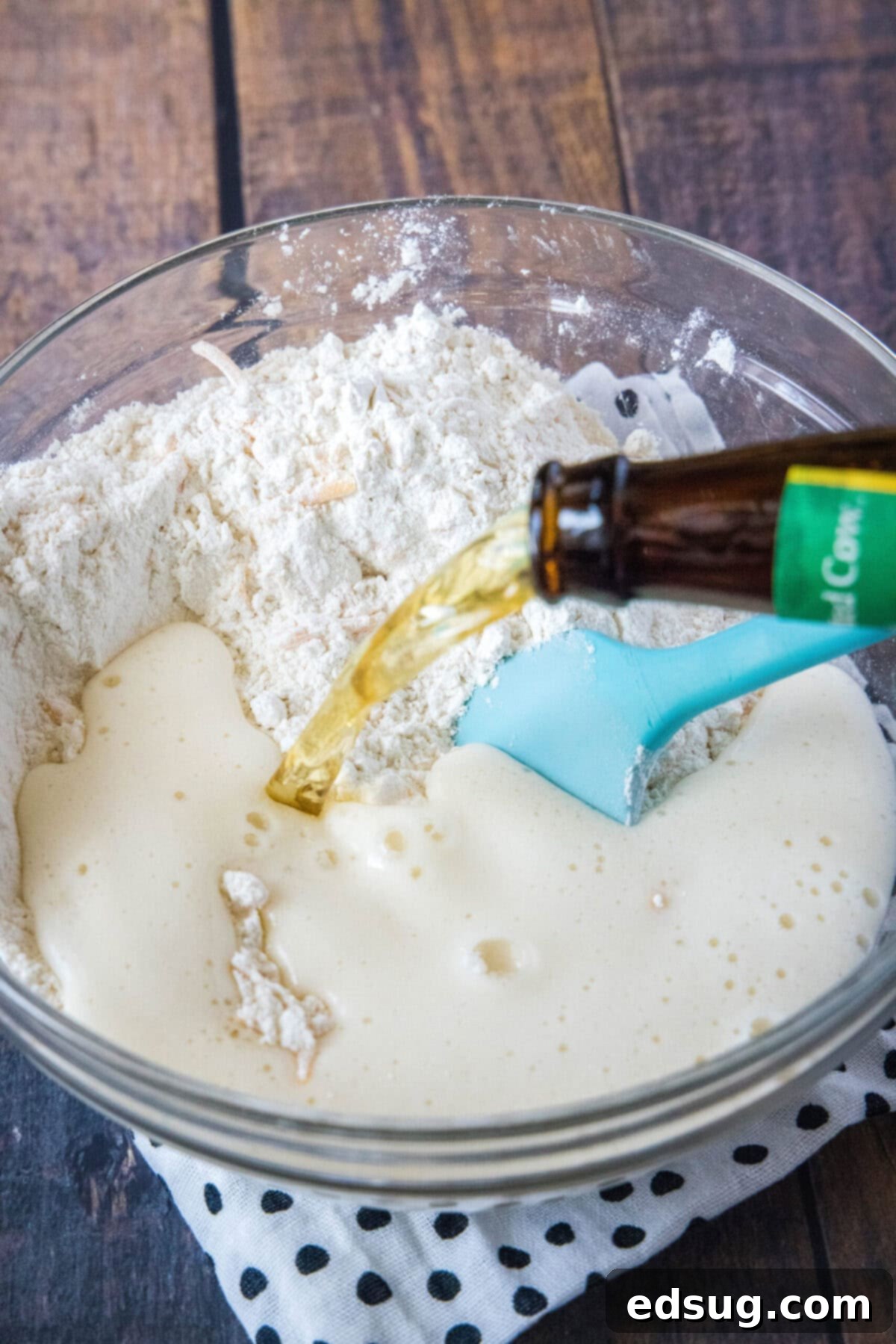 A refreshing bottle of beer being poured steadily into a bowl containing the mixed dry ingredients and cheese, with a rubber spatula beginning the gentle folding process to combine everything.