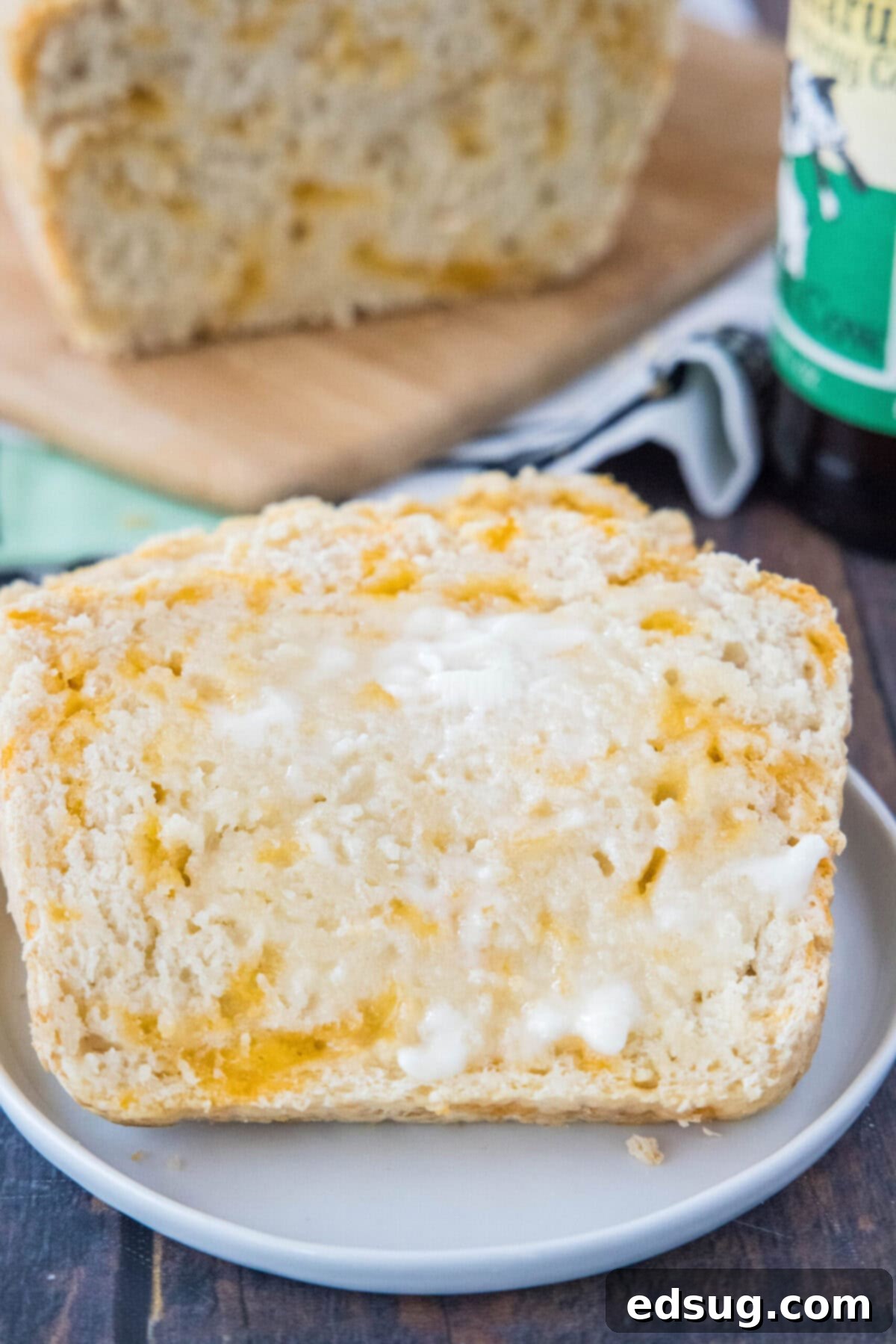 A perfectly baked, golden-crusted slice of cheddar beer bread, generously slathered with butter and ready to eat, artfully leaning against the main loaf in the background, inviting a delicious bite.