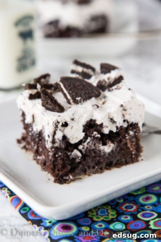 Delicious slice of Oreo Poke Cake on a white plate, showing layers of cake, condensed milk, pudding, and whipped cream with crushed Oreos.
