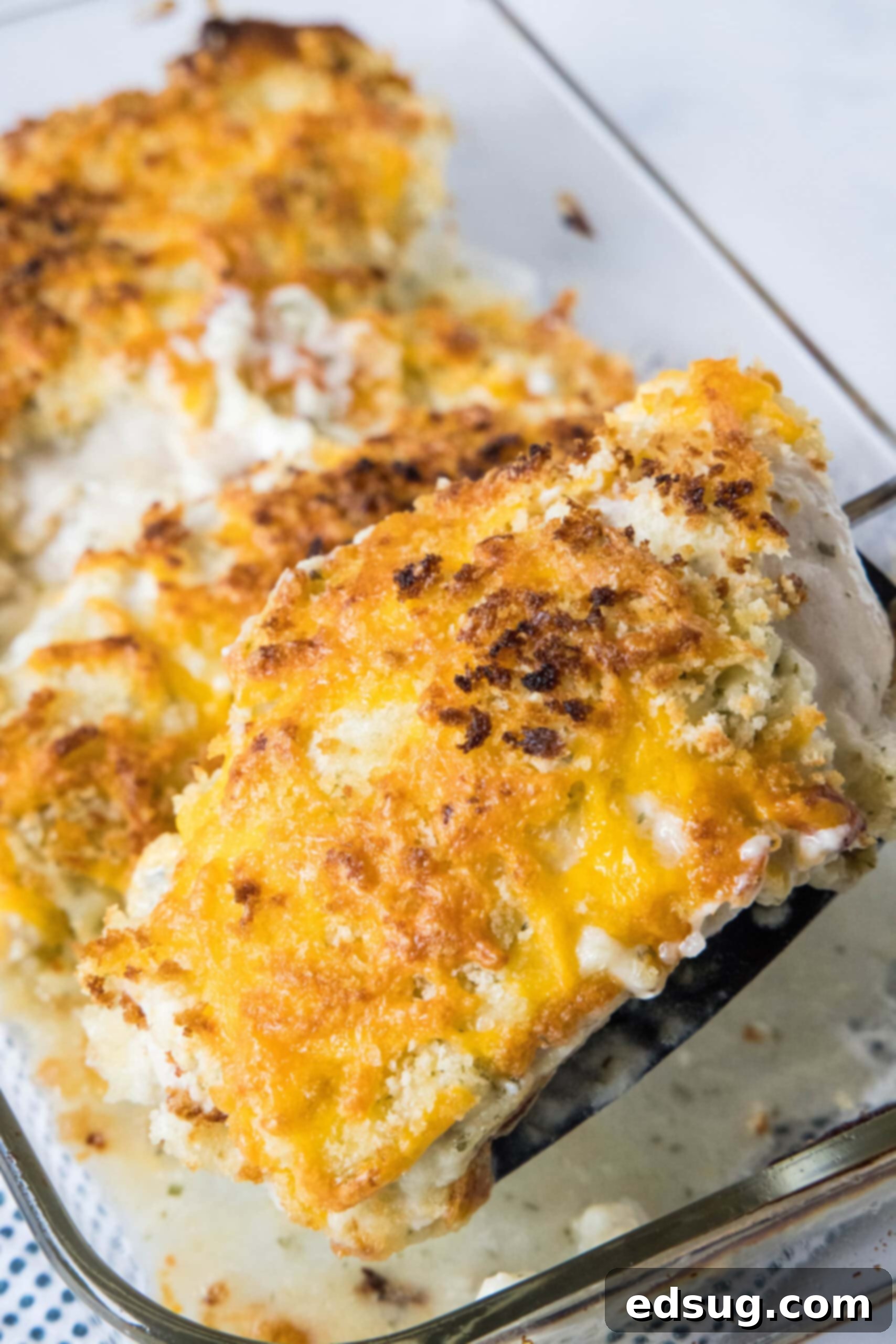 A spatula lifting a cheesy ranch chicken breast from a glass baking dish.
