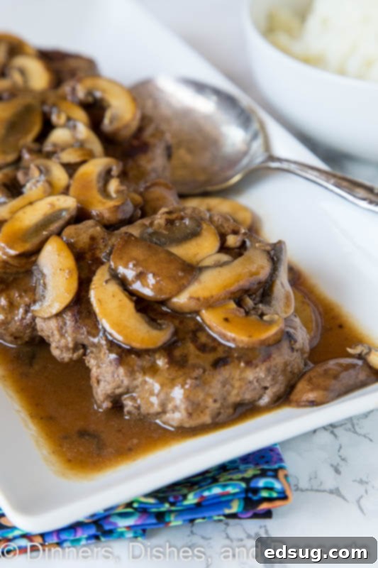Close-up of a plate with two hamburger steaks generously covered in a creamy, dark mushroom gravy, accompanied by a side of mashed potatoes.