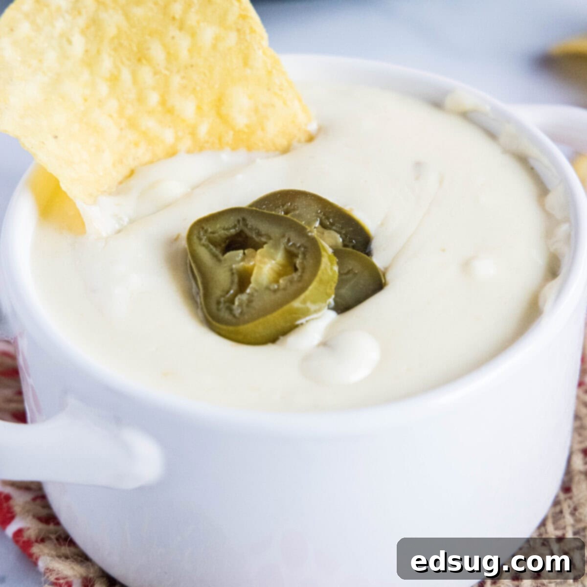 Kickoff Comfort Food 16 Close up of a bowl of creamy white queso dip, topped with pickled jalapeños, with a chip sticking in it