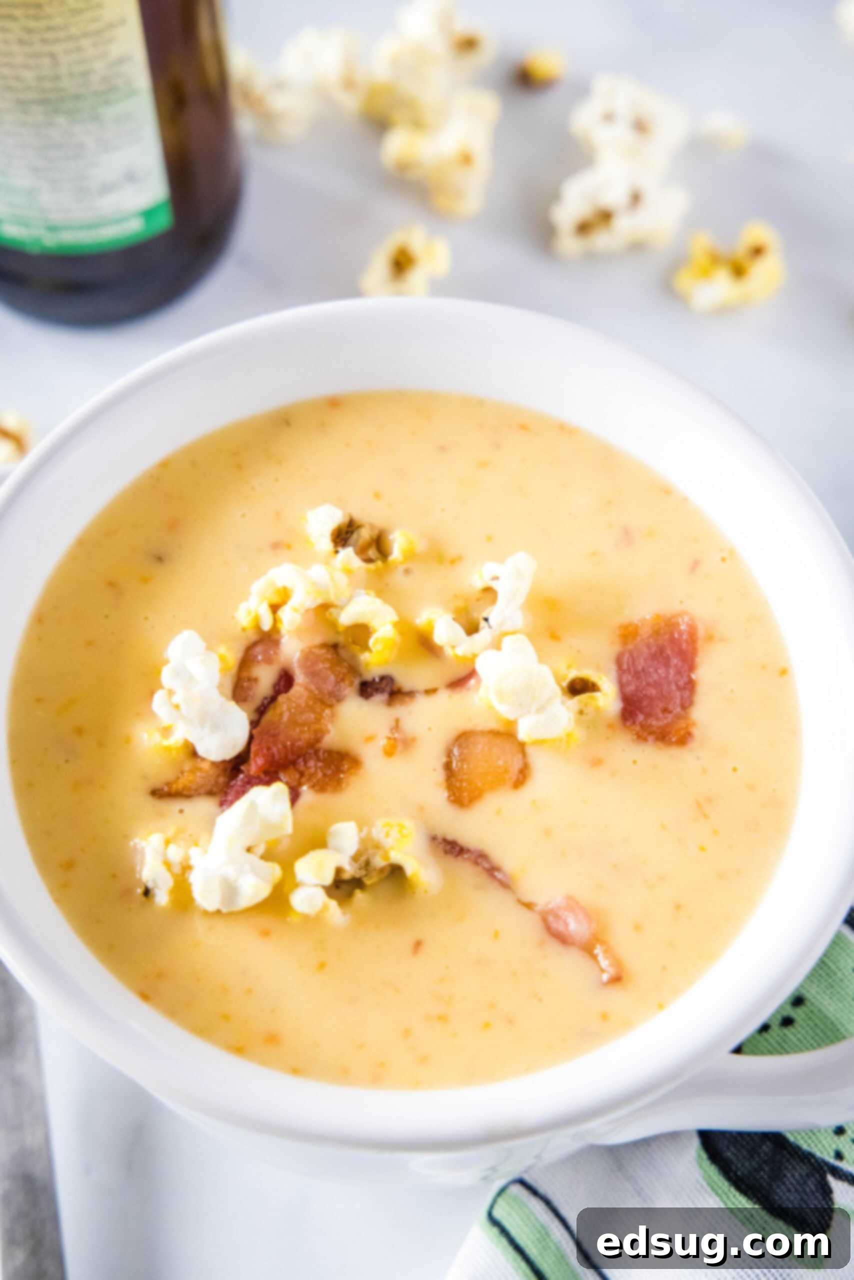 A bowl of creamy beer cheese soup, generously topped with crispy popcorn and savory bacon bits. In the background, a bottle of beer and some scattered popcorn enhance the cozy, indulgent scene.
