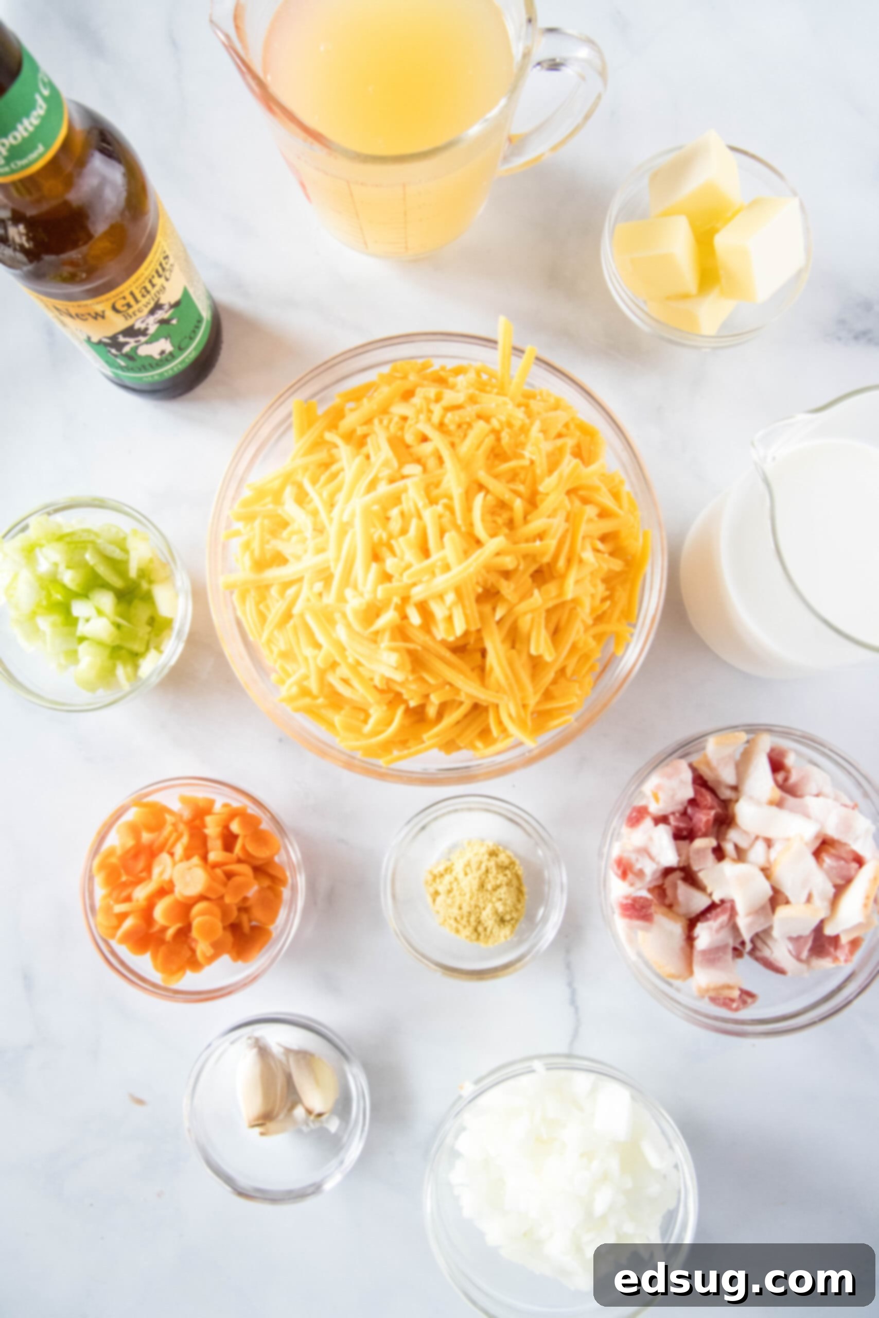 An overhead shot showcasing the fresh ingredients required for making beer cheese soup. Prominently displayed are a bottle of amber beer, a bowl of soft butter, a mound of grated sharp cheddar cheese, a pitcher of creamy milk, a bowl of neatly diced celery, another bowl of vibrant diced carrots, a small container of dried mustard powder, whole garlic cloves, finely diced onions, and a bowl of chopped raw bacon.