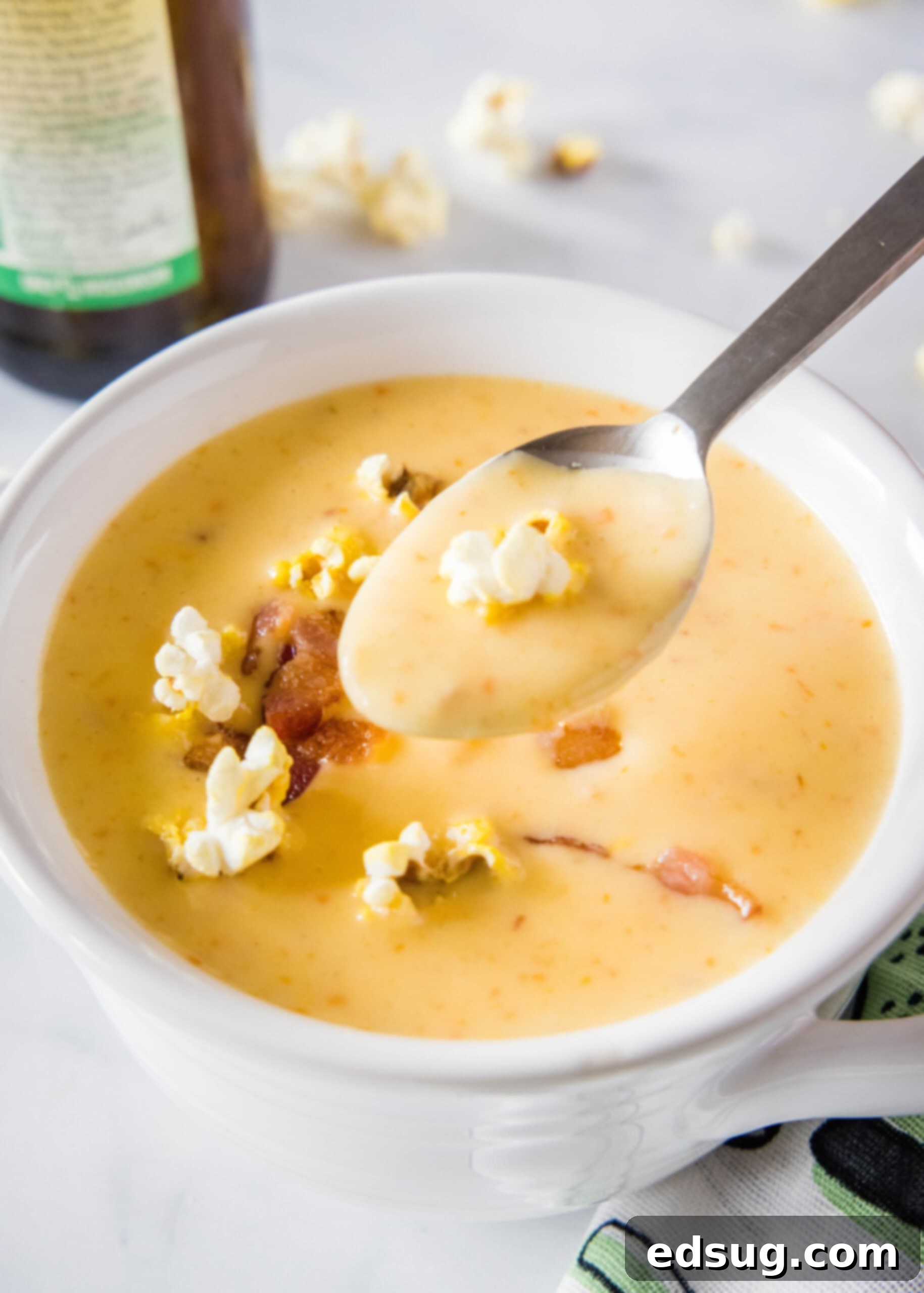 A close-up view of a bowl of creamy beer cheese soup, garnished with crispy bacon and fluffy popcorn. A spoon dips into the soup, scooping up a rich, cheesy bite. A bottle of beer stands blurred in the background, completing the inviting scene.