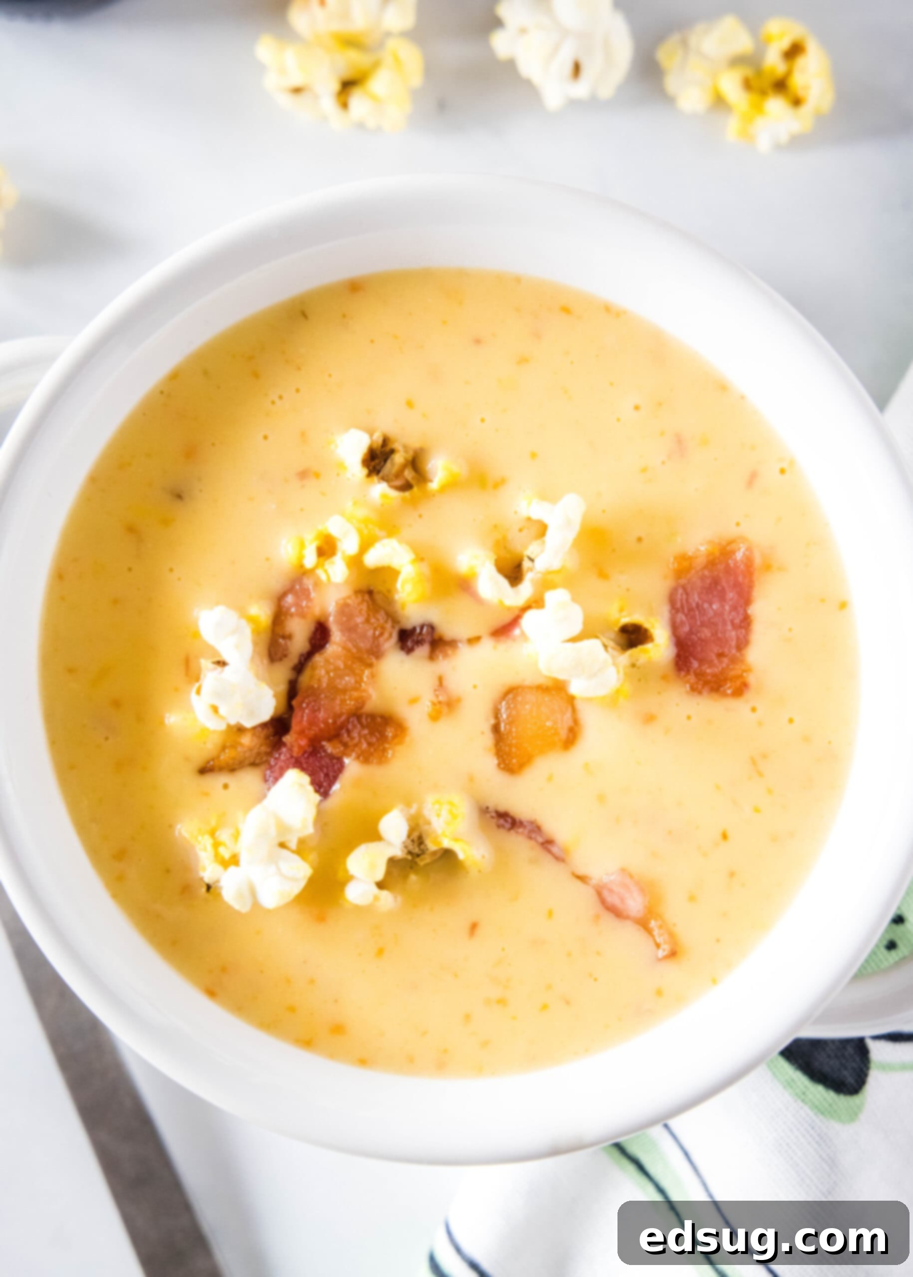 An inviting overhead view of a bowl of creamy beer cheese soup, adorned with golden popcorn and crispy bacon bits. A separate small bowl of popcorn sits on the side, ready for extra snacking.