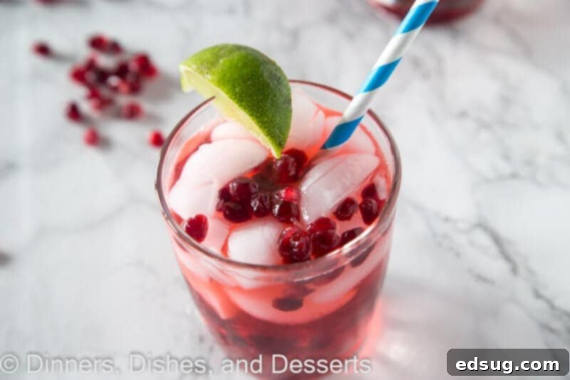Pomegranate vodka gimlet in a clear glass with a straw, garnished with lime and pomegranate seeds, showing its inviting red hue.