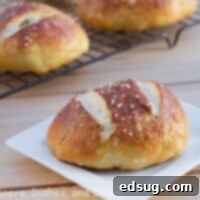 Soft Pretzel Buns 2 Close-up of a single golden-brown pretzel roll on a plate, with more rolls visible on a cooling rack in the background.