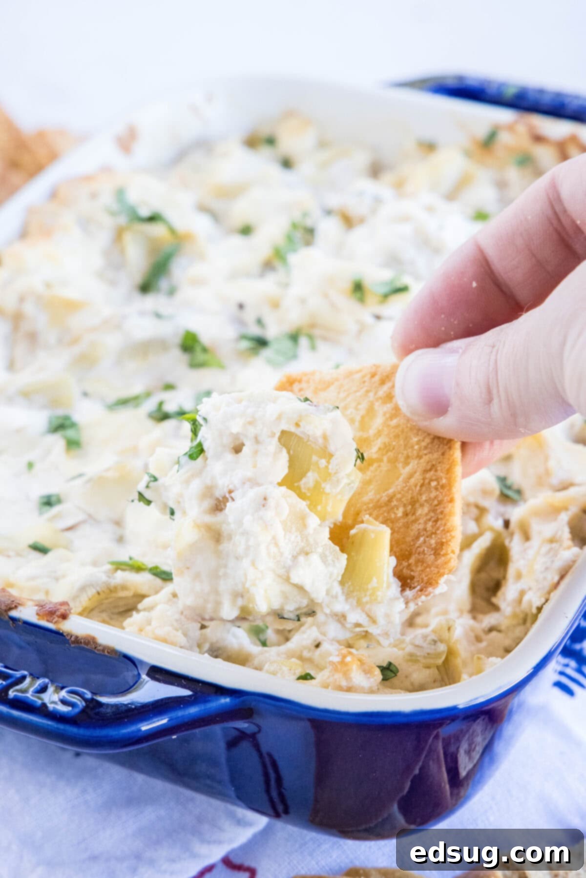 A tortilla chip scooping a generous portion of cheesy artichoke dip from a pan.