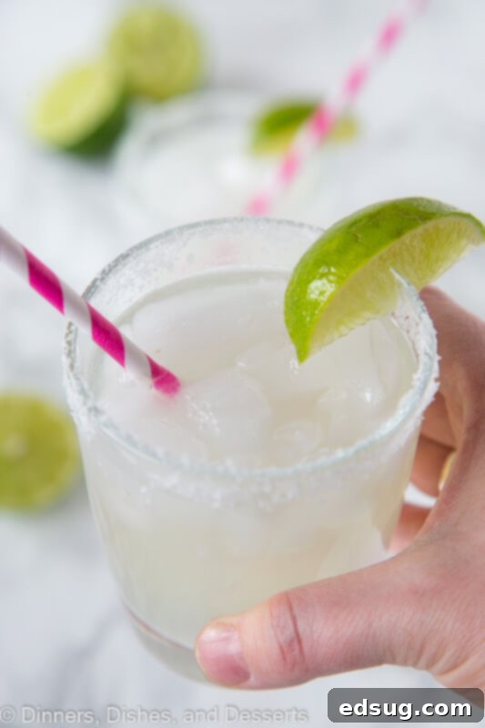 The Quintessential Lime Margarita, On the Rocks 3 A hand holds a classic lime margarita glass, garnished with a lime wedge and a pink straw, set against a bright, inviting background.