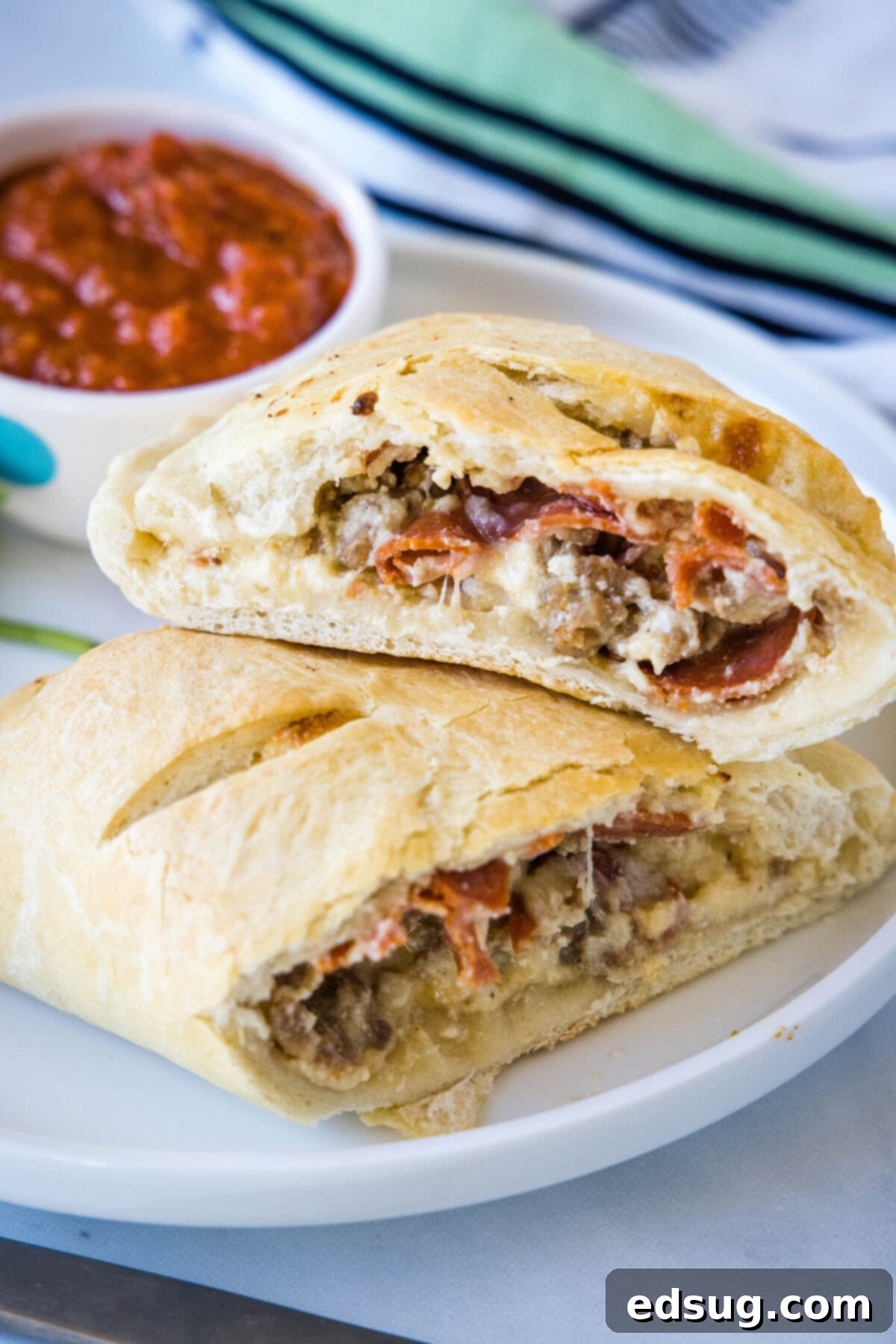 Folded Pizza Pockets 9 Two homemade calzones cut in half on a plate, revealing the sausage, pepperoni, and cheese filling, with a small bowl of pizza sauce for dipping.