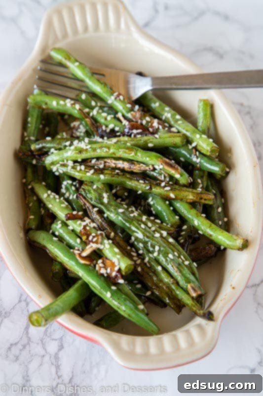 A serving dish filled with perfectly roasted green beans, glistening with sesame oil and soy sauce, generously sprinkled with toasted sesame seeds, and a fork ready to serve.