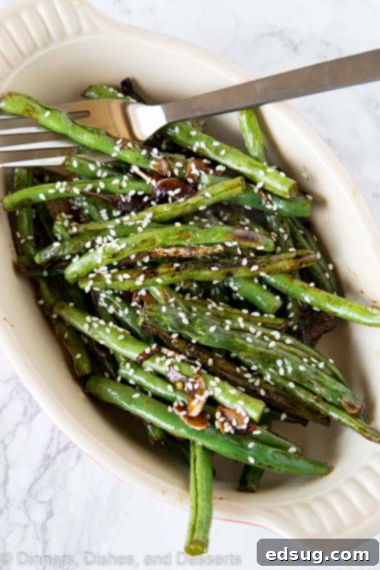 A close-up of roasted green beans being tossed with sesame seeds in a white bowl, ready for serving.