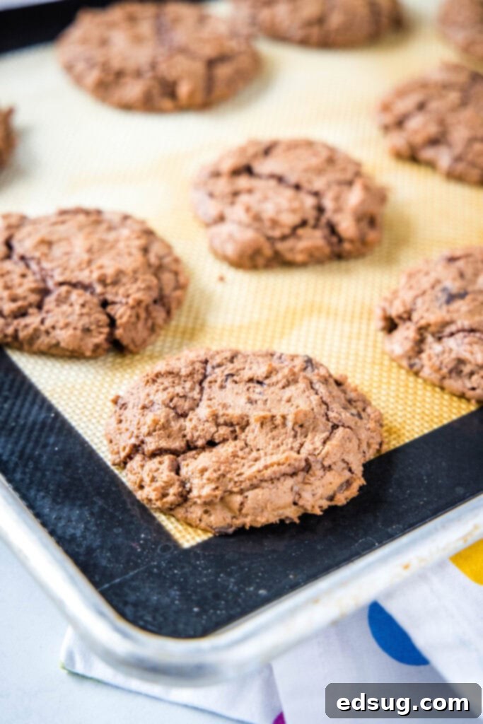 Decadent Chocolate Fudge Cookies 5 Freshly baked brownie cookies cooling on a baking sheet, showcasing their glossy, enticing crackly tops and soft centers.