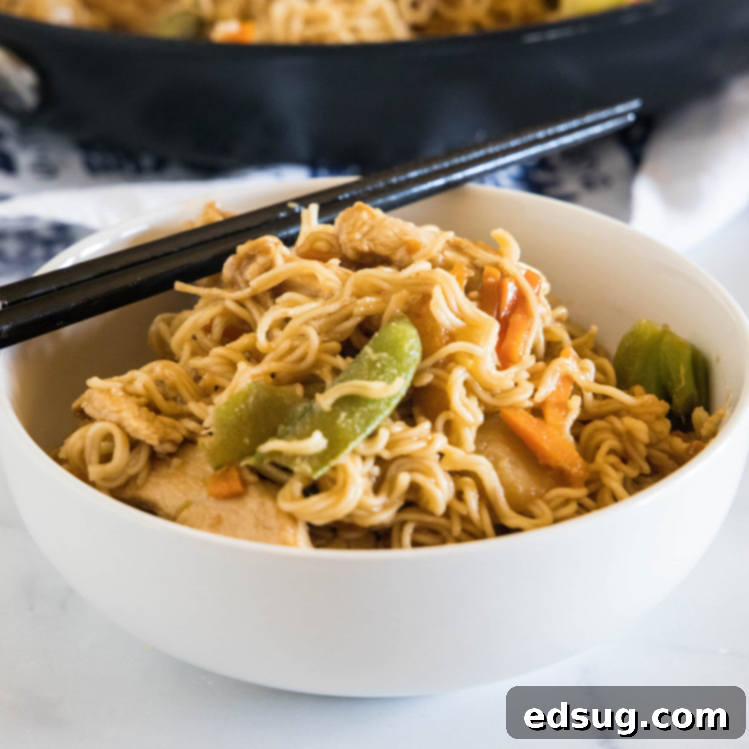 Homemade Asian Outshining the Takeout 10 Ramen stir fry in a white bowl with a set of chopsticks resting on the rim of the bowl.
