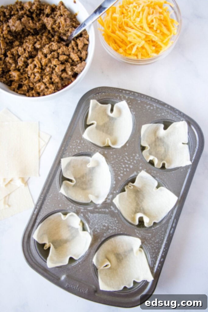 A 12-cup muffin tin meticulously lined with delicate wonton wrappers, positioned next to bowls of seasoned ground beef and shredded cheddar cheese, prepared for filling.
