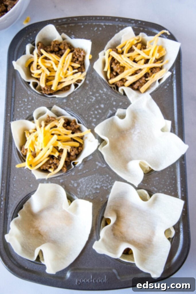 Several partially filled taco cups shown in a muffin tin, displaying the first layer of wonton wrappers, seasoned ground beef, and melted cheese, ready for the next layer.