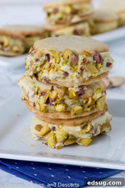 Pistachio ice cream sandwiches stacked on a plate, showing creamy pistachio ice cream between two golden sugar cookies, coated with chopped pistachios