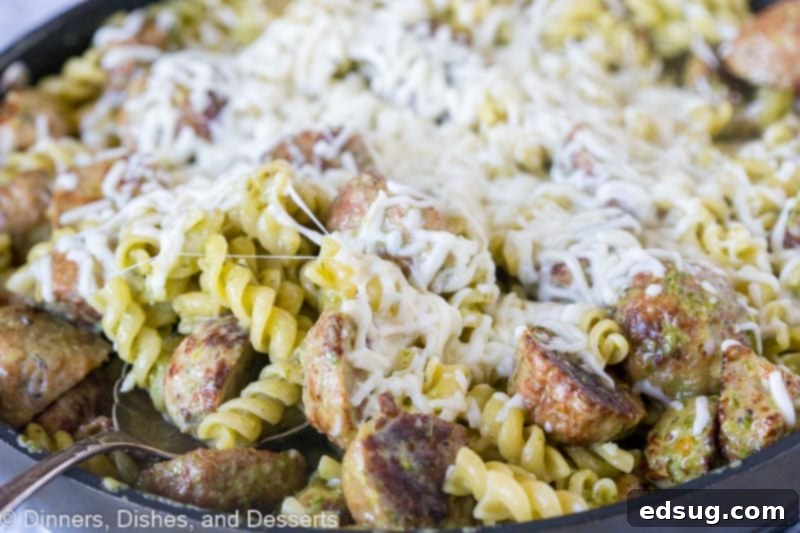 Skillet filled with creamy pesto pasta and meatballs, with a layer of cheese melting on top.