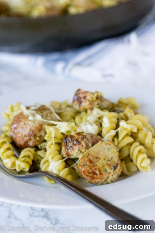 A fork taking a bite of creamy pesto pasta with a meatball, highlighting the melted cheese.