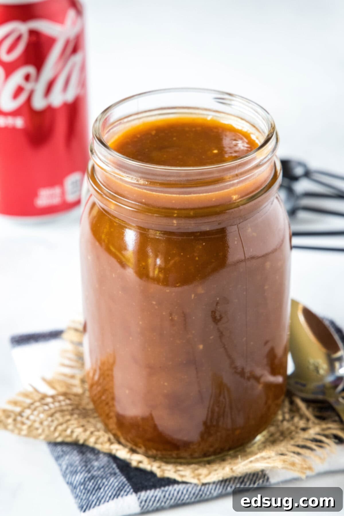 A jar of homemade Coca-Cola BBQ sauce with a can of Coke in the background, highlighting its sweet, spicy, and sticky qualities.