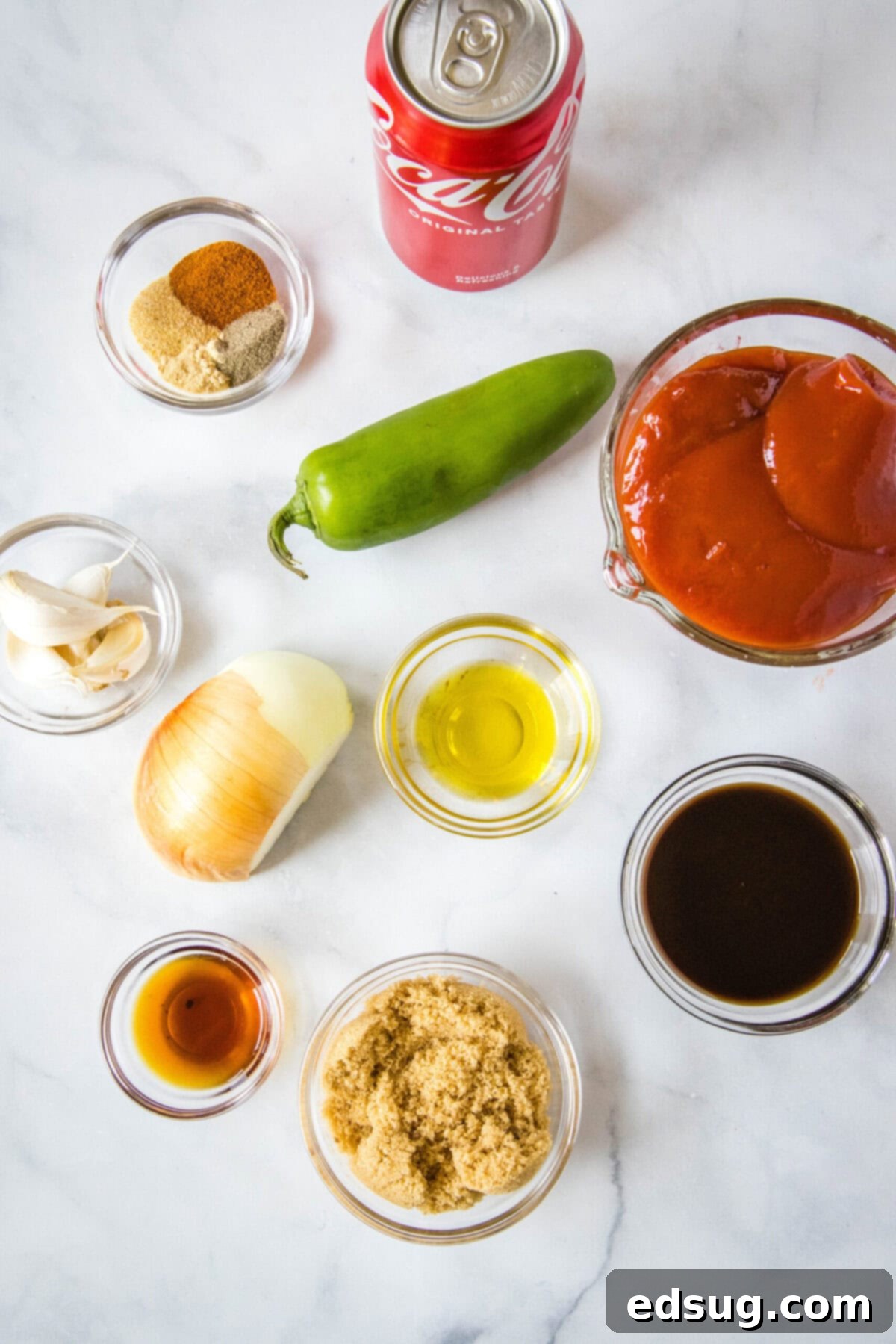 Overhead view of all the fresh ingredients needed for homemade Coca-Cola BBQ sauce, including a can of Coke, ketchup, jalapeño, garlic, oil, onion, brown sugar, Worcestershire sauce, liquid smoke, and various spices.