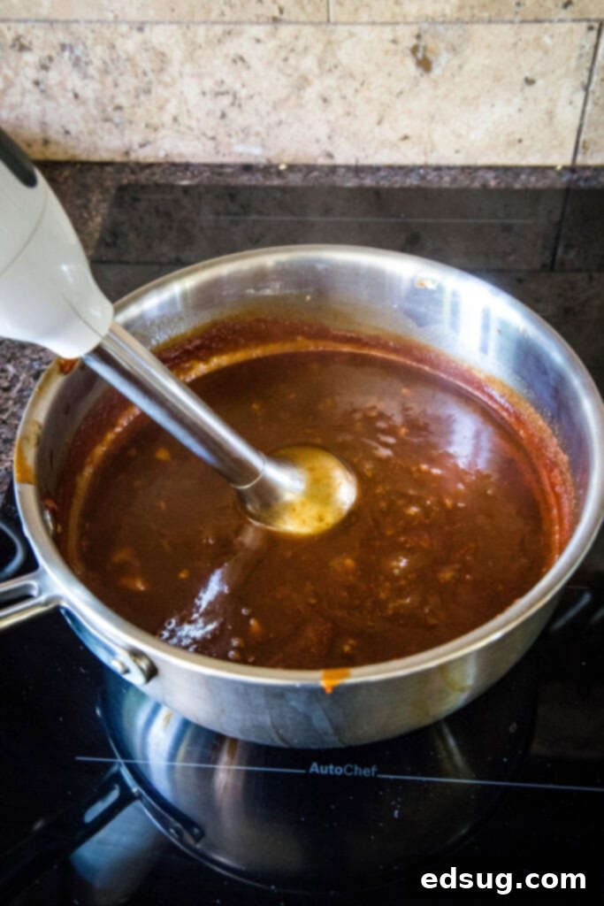 An immersion blender smoothly pureeing the Coca-Cola BBQ sauce directly in the pot to achieve a perfect consistency.