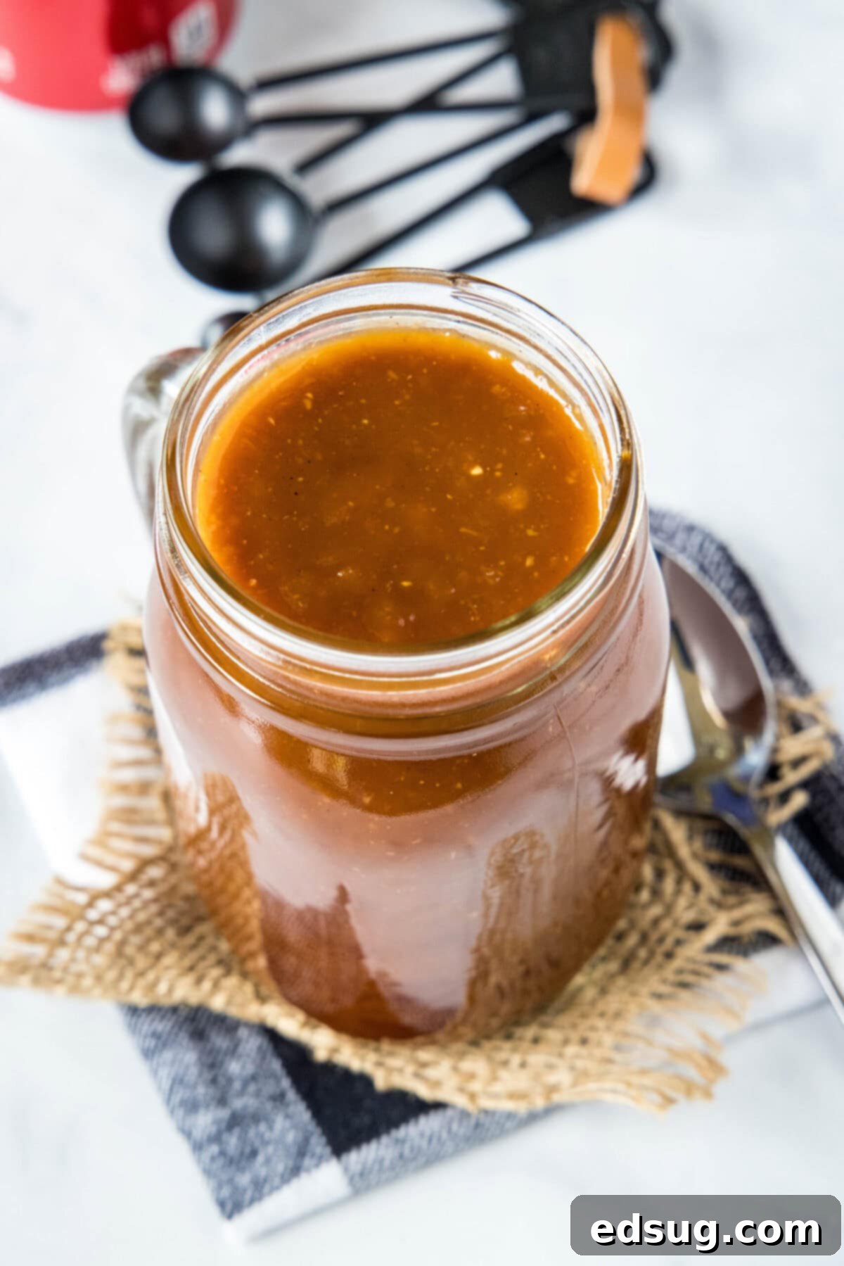 Overhead view of a jar of homemade Coca-Cola BBQ sauce, with a spoon and measuring spoons, emphasizing its homemade goodness and potential for storage.